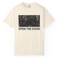 Load image into Gallery viewer, An off-white short-sleeve t-shirt with a graphic print of three black cats with glowing orange eyes and the text 'OPEN THE DOOR' underneath, displayed on a clean white background.