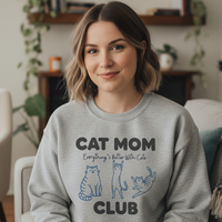 Load image into Gallery viewer, Woman wearing a gray sweatshirt with &#39;Cat Mom Club&#39; text and cat illustrations in a home setting.
