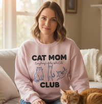 Load image into Gallery viewer, Woman wearing a pink sweatshirt with &#39;Cat Mom Club&#39; text and cat illustrations, sitting on a couch with a cat.

